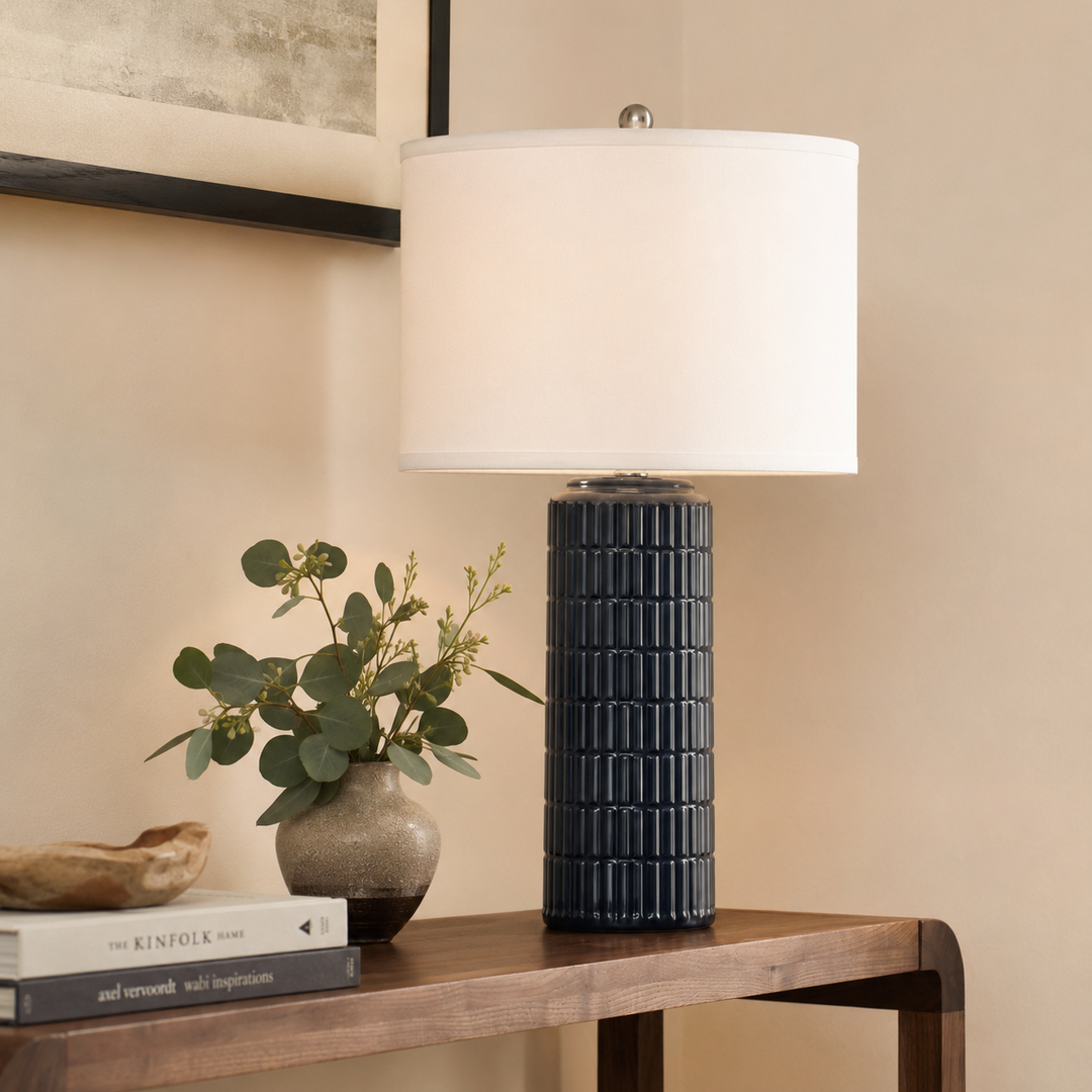 A table lamp with a white drum shade and a dark navy ribbed ceramic base, styled on a wooden console table next to a vase of greenery.